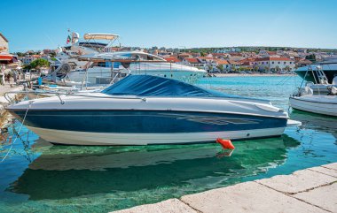 Parlak mavi bir gökyüzünün altında kıyı binaları ve diğer teknelerle berrak, berrak bir marinaya parlak bir motorlu tekne yanaştı..