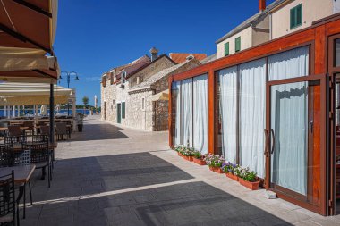 Huzurlu deniz kenarı restoranı, modern ahşap masalar ve büyük şemsiyelerin altında deniz manzaralı bir gezinti alanı boyunca sıralanmış sandalyeler..