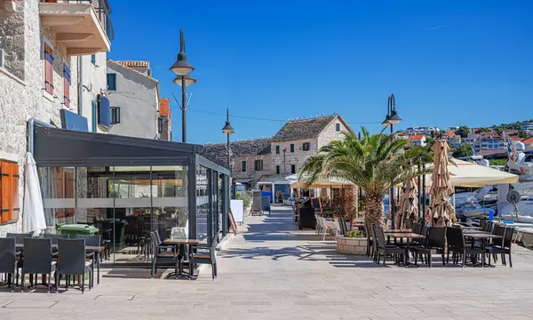 Beyaz hasır mobilyaları, çizgili yastıkları ve kapalı şemsiyeleri olan güneşli bir açık kafe terası. Akdeniz 'de palmiye ağaçları ve çiçek ekicileri olan bir plazada geçiyor..