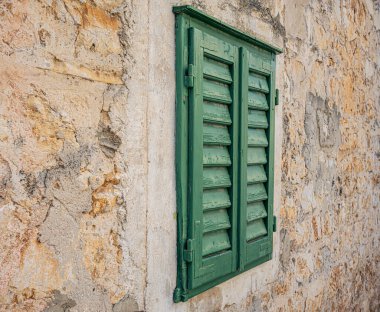 Yıpranmış yeşil ahşap kepenk, desenli, açık renkli taş bir duvara dayanıyor ve geleneksel Akdeniz mimarisinin kırsal çekiciliğini sergiliyor..