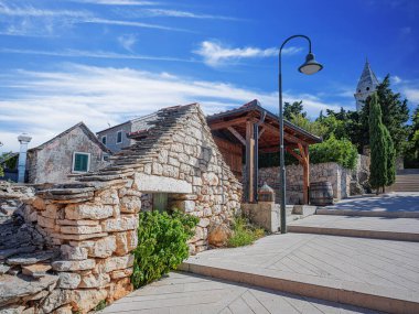 Hırvatistan 'ın Primosten kentindeki günışığı sokak sahnesinde geleneksel taş mimari, belirgin bir çan kulesi ve ince bulutlu dinamik mavi gökyüzü altında yemyeşil bir Akdeniz kulesi yer alıyor..