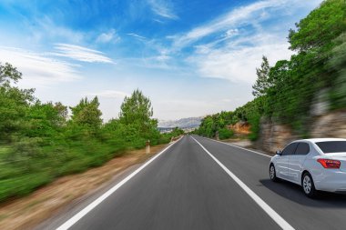 Bulanık bir otoyolda ilerleyen beyaz bir sedan. Bulanık bir mavi gökyüzünün altında yemyeşil bir kanat var. Resimli bir manzarada dinamik bir yolculuk olduğunu gösteriyor..
