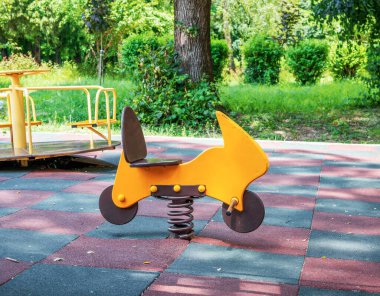 Motosiklet şeklinde parlak renkli sarı ve kahverengi bir yay binicisi güneşli arka planda yeşil ağaçlar ve çimlerle kaplı kareli kauçuk bir oyun alanı yüzeyinde duruyor..
