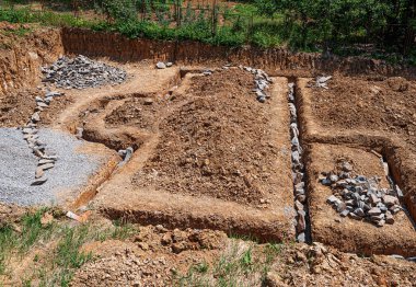 Açık hava inşaat sahasında kayalar ve çakıllarla dolu yeni kazılmış çukurlar görülüyor. Temiz güneş ışığı altındaki bir binanın ilk aşamalarını işaret ediyor..