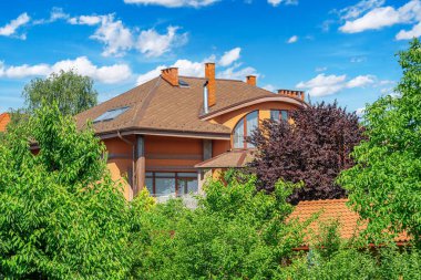 Kahverengi kiremit çatılı modern banliyö evi. Etrafı yemyeşil ağaçlarla çevrili. Parlak mavi gökyüzü altında dağınık bulutlar..