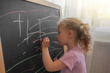 Pembe tişörtlü genç bir kız, karatahtaya yoğunlaşarak renkli çizgiler çiziyor, erken çocukluk eğitimini ve yaratıcı ifadesini kapalı alanlarda vurguluyor..