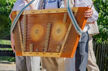 Yakın plan çekimde geleneksel kıyafetli bir müzisyenin açık havada santur çaldığı görülüyor..