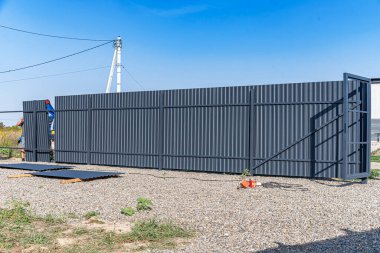 Kısmen döşenmiş, kapısı açık, çakılla kaplı bir arazide inşaat malzemeleri ve aletleri etrafa saçılmış vaziyette bulunan metal bir çit.