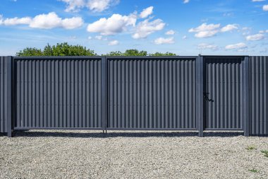 Modern gri kıvrımlı metal bir çit, yayaya ait bir kapı, parlak mavi bir gökyüzünün altında, dağınık bulutlu, çakıl taşı bir yüzey üzerinde duruyor..