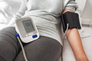 Bir kişi evde otururken dijital monitörle kan basıncını ölçer ve kişisel sağlık ve tıbbi bakım konularına odaklanır..