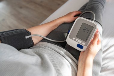 Bir kişi evde otururken dijital monitörle kan basıncını ölçer ve kişisel sağlık ve tıbbi bakım konularına odaklanır..