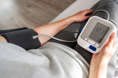 Bir kişi evde otururken dijital monitörle kan basıncını ölçer ve kişisel sağlık ve tıbbi bakım konularına odaklanır..