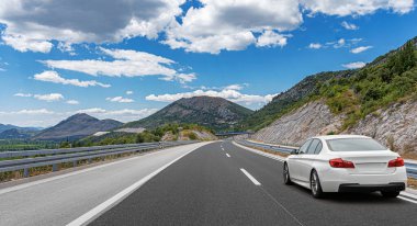 Bereketli bir yeşil ve kayalık arazi ile çevrili, canlı bir mavi gökyüzü altında dolambaçlı bir dağ otoyolu boyunca sürüş beyaz araba manzaralı bir yol konsepti için.