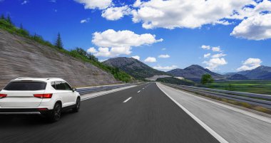Beyaz SUV, parlak mavi bir gökyüzü altında, dağ yolunda hızla ilerliyor. Etrafı kayalık yamaçlar ve yeşil vadilerle çevrili. Özgürlüğü ve seyahati simgeliyor..