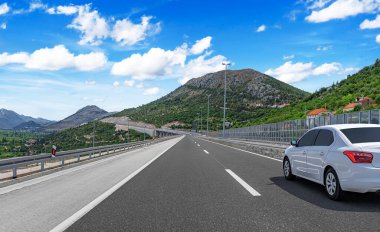 Beyaz bir araba, yeşil tepeler ve manzaralı manzarayla çevrili, açık mavi bir gökyüzü altında pürüzsüz bir dağ yolu boyunca sürüyor..