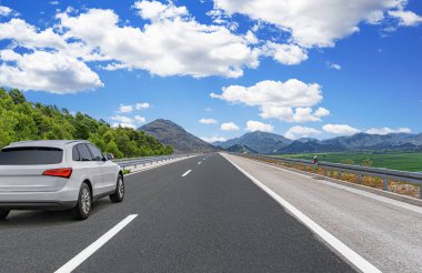 Beyaz SUV, parlak mavi bir gökyüzü altında, dağ yolunda hızla ilerliyor. Etrafı kayalık yamaçlar ve yeşil vadilerle çevrili. Özgürlüğü ve seyahati simgeliyor..