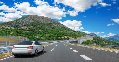 Bereketli bir yeşil ve kayalık arazi ile çevrili, canlı bir mavi gökyüzü altında dolambaçlı bir dağ otoyolu boyunca sürüş beyaz araba manzaralı bir yol konsepti için.