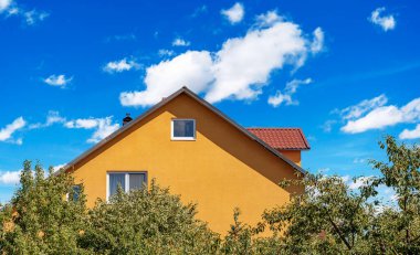 Kırmızı çatılı canlı sarı bir ev parlak mavi bir gökyüzüne karşı duruyor. Kabarık beyaz bulutlar, kısmen önplanda yeşil ağaçlar tarafından gizleniyor..
