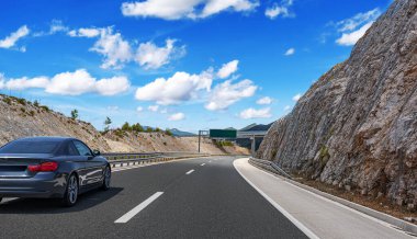 Modern siyah coupe, kayalık bir yamacın yanında, bulutlu, parlak mavi bir gökyüzünün altında, manzaralı bir otoyolda sürüyor..