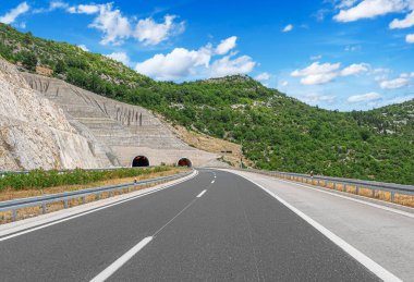 Uzun, dolambaçlı bir otoyol, açık mavi gökyüzünün altında, doğal bir manzarada kabarık beyaz bulutlar, seyahat ve altyapısı olan yemyeşil dağlara oyulmuş iki tünele götürür..