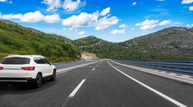 Beyaz SUV, parlak mavi bir gökyüzü altında, dağ yolunda hızla ilerliyor. Etrafı kayalık yamaçlar ve yeşil vadilerle çevrili. Özgürlüğü ve seyahati simgeliyor..