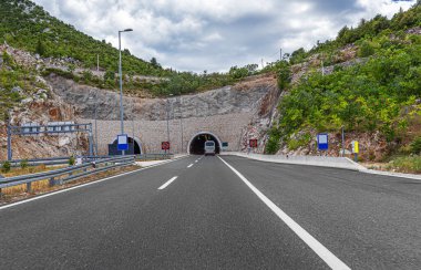 Modern bir otoyol, bulutlu bir gökyüzünün altında yeşil, kayalık dağlarla çevrili bir yol tünelinin girişine yaklaşıyor..