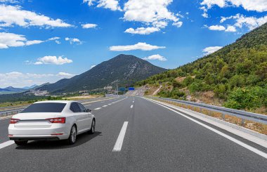 Beyaz bir araba modern bir otoyolda gidiyor. Manzaralı bir dağ manzarası boyunca. Parlak mavi bir gökyüzünün altında dağınık bulutlarla..