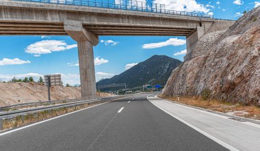 Geniş beton bir üst geçidin altında, açık mavi gökyüzü ve dağınık bulutlarla modern bir otoyol, dağlık bir arazide esiyor..