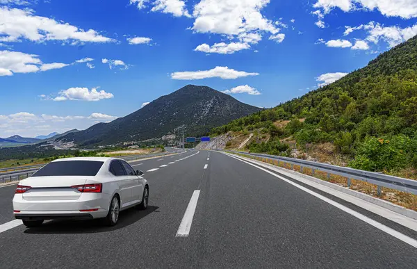 Beyaz bir araba modern bir otoyolda gidiyor. Manzaralı bir dağ manzarası boyunca. Parlak mavi bir gökyüzünün altında dağınık bulutlarla..