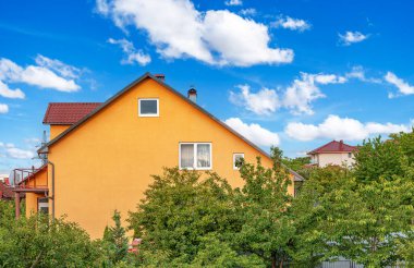 Kırmızı çatılı canlı sarı bir ev parlak mavi bir gökyüzüne karşı duruyor. Kabarık beyaz bulutlar, kısmen önplanda yeşil ağaçlar tarafından gizleniyor..