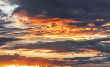 Parlayan turuncu bulutlarla dolu dramatik bir akşam gökyüzü batan güneş tarafından karanlık ve kasvetli bir zemin üzerine aydınlatılıyor..