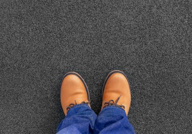 Kahverengi deri çizme ve mavi kot giyen, koyu desenli kaldırımda dikilen, sıradan moda ve ayakkabıları temsil eden bir insanın tepeden aşağı bakışı..