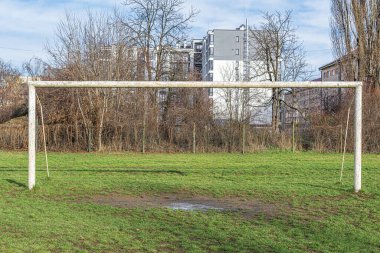 Arka planda yapraksız ağaçlar ve konut binaları olan çimenli bir şehir sahasında paslı bir kale direği..