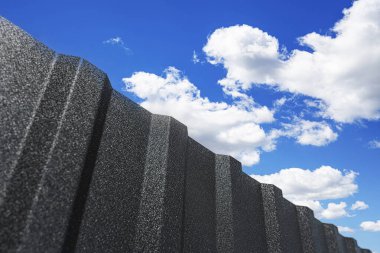 Koyu, dokulu metal bir çitin alçak açılı görüntüsü, kabarık beyaz bulutlu, canlı mavi bir gökyüzüne karşı..