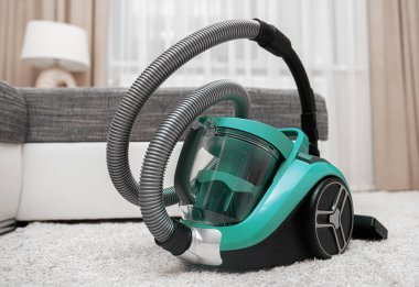 Şeffaf bir toz kabı olan modern yeşil bir elektrikli süpürge rahat bir oturma odasında yumuşak beyaz bir halının üzerinde duruyor..