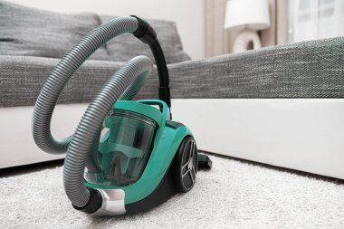 Şeffaf bir toz kabı olan modern yeşil bir elektrikli süpürge rahat bir oturma odasında yumuşak beyaz bir halının üzerinde duruyor..