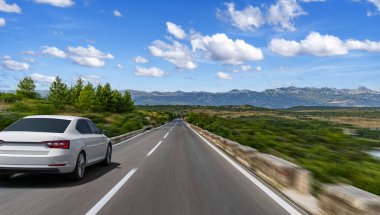 Açık bir otoyolda hızla ilerleyen beyaz bir araba manzaralı bir dağ manzarası boyunca dağınık bulutlu açık mavi bir gökyüzünün altında..
