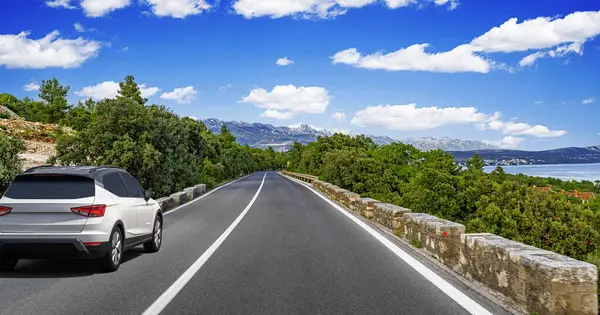Beyaz bir SUV, bereketli yeşilliklerle çevrili, uzak dağları ve parlak mavi gökyüzünün altında ışıl ışıl parlayan bir denizi olan manzaralı bir sahil yolunda ilerliyor..
