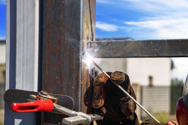 Parlak mavi gökyüzünün altında görünür kıvılcımlar, duman ve koruyucu eldivenlerle metal kaynak yapan bir işçinin yakın çekimi..