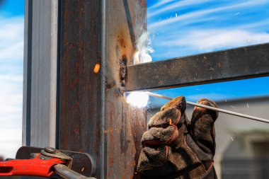 Parlak mavi gökyüzünün altında görünür kıvılcımlar, duman ve koruyucu eldivenlerle metal kaynak yapan bir işçinin yakın çekimi..