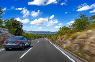 Boş bir dağ yolunda giden siyah lüks bir araba. Etrafı yemyeşil, parlak mavi bir gökyüzünün altında dağınık bulutlarla çevrili..
