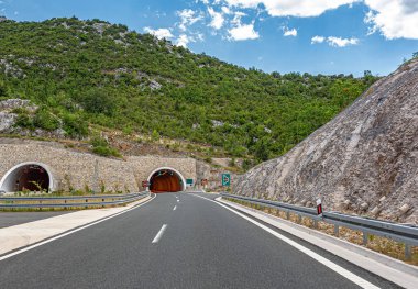 Parçalı bulutlu bir yaz gökyüzünün altında kayalık yamaçlar ve yemyeşil yemyeşil ile çevrili ikiz dağ tünellerine doğru modern bir otoyol kıvrılır..
