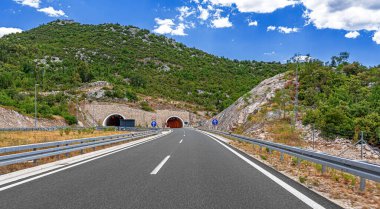 Parçalı bulutlu bir yaz gökyüzünün altında kayalık yamaçlar ve yemyeşil yemyeşil ile çevrili ikiz dağ tünellerine doğru modern bir otoyol kıvrılır..