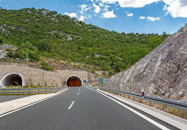 Parçalı bulutlu bir yaz gökyüzünün altında kayalık yamaçlar ve yemyeşil yemyeşil ile çevrili ikiz dağ tünellerine doğru modern bir otoyol kıvrılır..
