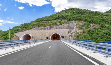 Parçalı bulutlu bir yaz gökyüzünün altında kayalık yamaçlar ve yemyeşil yemyeşil ile çevrili ikiz dağ tünellerine doğru modern bir otoyol kıvrılır..