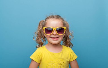 Sarı kıvırcık saçlı, sarı gözlüklü, atölyede mavi bir arka plana gülümseyen neşeli, genç bir kız bebek..