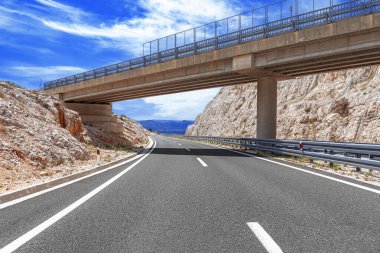 Modern bir beton köprü kıvrımlı bir otoyol üzerinde uzanır. Etrafı kayalık tepelerle çevrilidir. Parlak mavi bir gökyüzünün altında dağınık bulutlarla..
