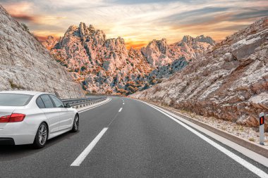 Beyaz bir sedan, kayalık uçurumlar ve dramatik dağlarla çevrili renkli bir günbatımı gökyüzü altında virajlı bir otoyolda ilerliyor..