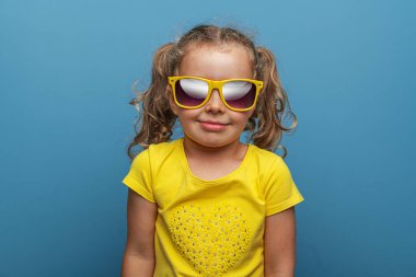 Sarı kıvırcık saçlı, sarı gözlüklü, atölyede mavi bir arka plana gülümseyen neşeli, genç bir kız bebek..