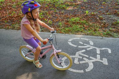 Renkli bir kask takan genç bir kız parktaki kaldırımlı bir yolda bisiklet sürüyor, dışarıda eğleniyor..